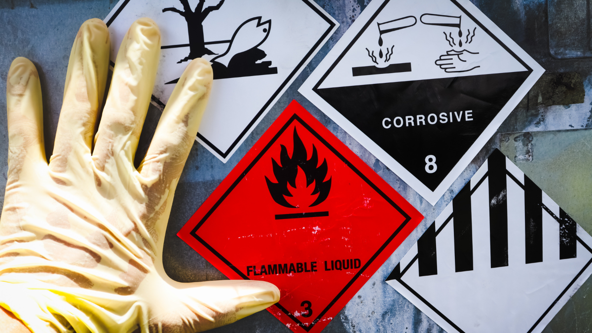 Safety gloves protecting against hazardous materials with warning labels for corrosive, flammable, and toxic substances in the background.