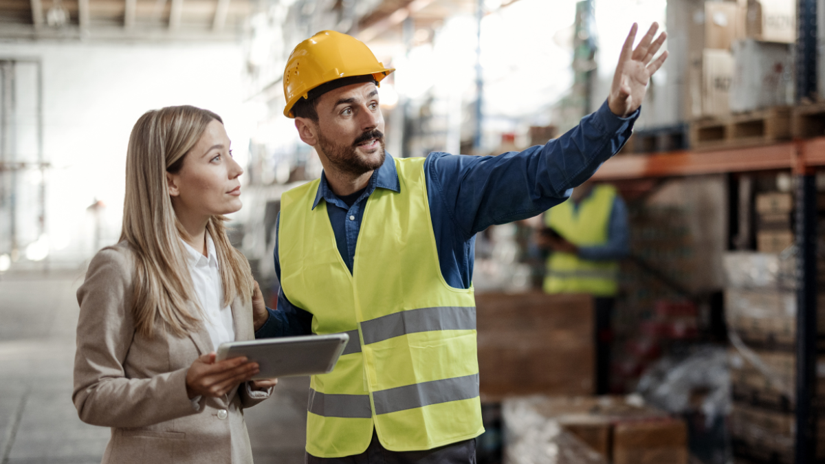 An OSH Coordinator in a safety vest and hard hat discussing workplace safety measures with a colleague holding a tablet in a warehouse setting.