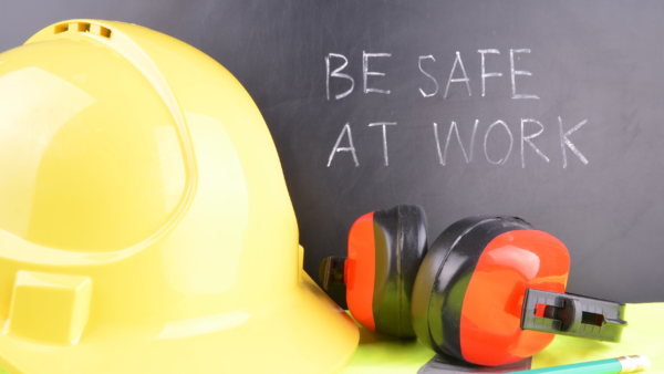 Yellow hard hat and protective ear muffs placed in front of a chalkboard with the message 'Be Safe at Work' written on it.