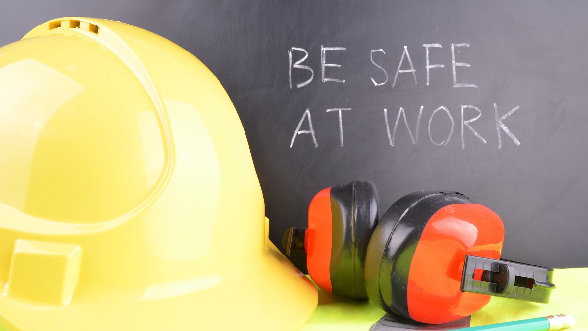 Yellow hard hat and protective ear muffs placed in front of a chalkboard with the message 'Be Safe at Work' written on it.