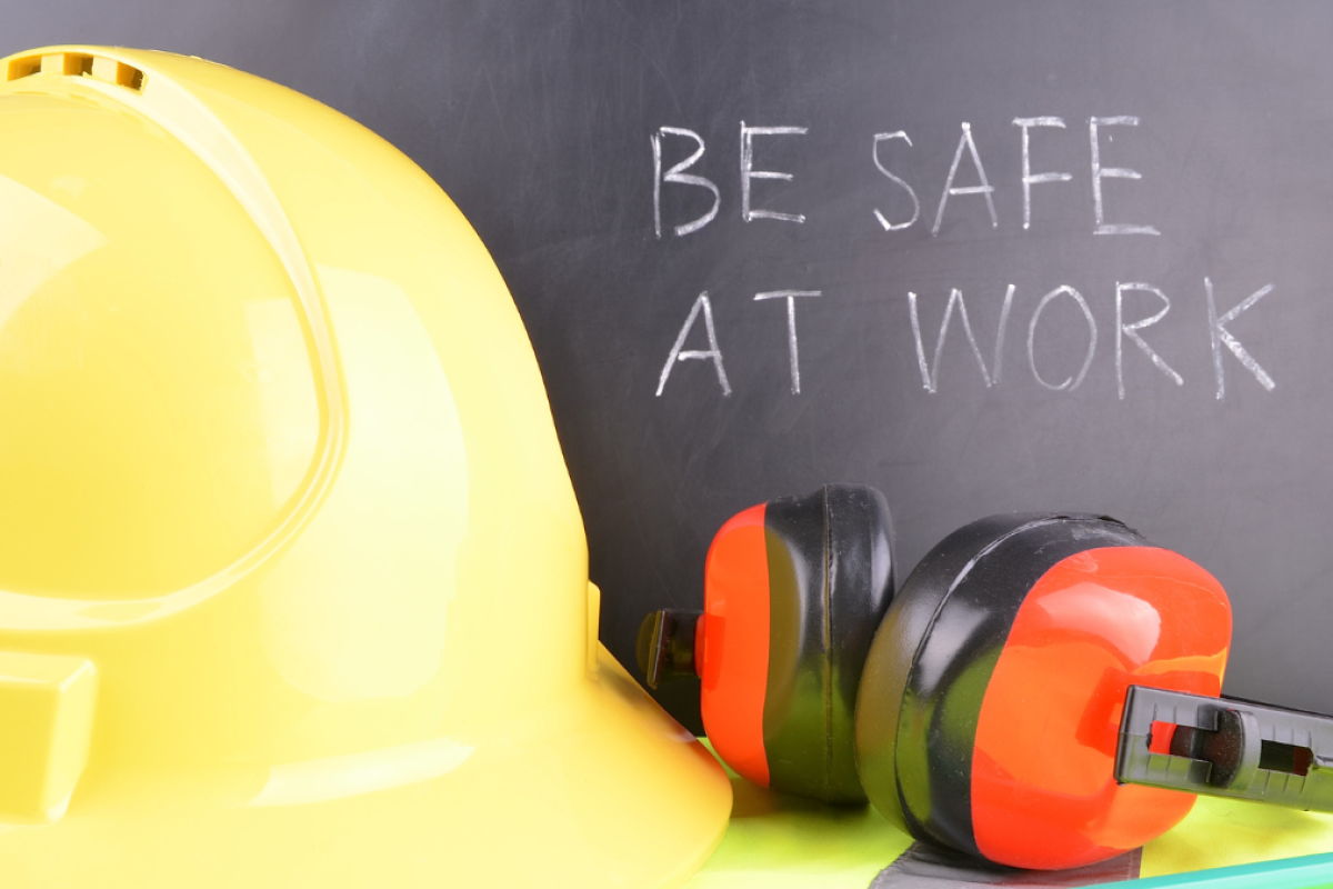 Yellow hard hat and protective ear muffs placed in front of a chalkboard with the message 'Be Safe at Work' written on it.